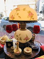Photo showing a floral hurricane lamp with pink rose designs, two 1-2 light lamps, four red glass goblets, a small floral vase, a small cup, and small plates, all displayed on a wooden surface near a window.