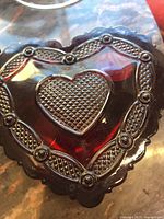 Top and angled views of a 4 inch heart-shaped red glass trinket box with textured heart pattern and decorative embossed border on the lid.