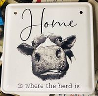 Close-up image of the square enamel sign showing the face of a black and white cow and the phrase 'Home is where the herd is'. The sign is white with black text and artwork, two holes for mounting.
