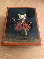 Top view of the closed wooden music box featuring a painted image of a girl in traditional attire holding a watering can with flower petals around her on dark background.