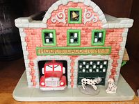 Front view of ceramic firehouse tealight candle holder showing the fire truck inside garage and two Dalmatian dogs in front.