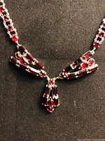 Close-up photo of the necklace pendant showing detailed red rhinestones arranged in a bow shape with dangling baguette cut stones on a silver plated setting.