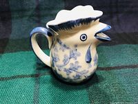 Front angle of blue and white ceramic creamer shaped like a rooster showing spout beak, comb rim, and painted floral details.