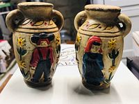 Pair of vintage ceramic folk art salt and pepper shakers depicting a man and woman with floral decorations on urn-shaped bodies with handles