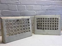 Photo showing front view of two vintage metal air vents placed side by side against a white brick wall. The vents have a honeycomb patterned grate and worn white paint.