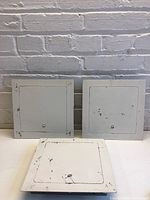 Three white square metal access panels with chipping paint arranged standing upright on a surface against a brick wall.