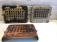 Three vintage decorative metal air vents showing different colors and wear levels arranged on white surface against a white brick wall.