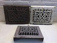 Three vintage decorative cast iron vents displayed against a white brick wall, showing varying paint colors and conditions.