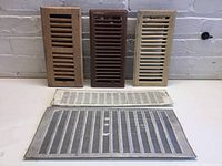 All 5 vents displayed together on a white surface against a brick wall. Three wooden vents are upright; the two metal vents lie flat in front.