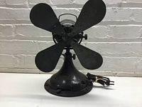 Front view of antique four-blade black metal fan on pedestal base with visible power cord.