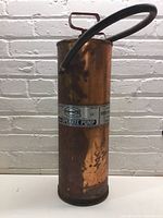 Full view of the antique copper fire extinguisher showing cylindrical body, handle, and hose against a white brick wall background.