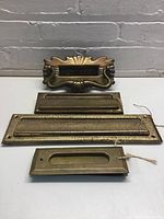 Four brass door mail slots with flaps laid out on a gray surface against a gray brick wall. Top piece is ornate Art Nouveau door knocker with attached screws. Below are three brass mail slots in varying sizes, one painted on one side, one showing rust spots, and a smaller slot with a flap.