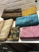 Photo shows six folded fabric remnants in multiple colors: brown ribbed, yellow textured, teal blue plush, dusty pink velvet-like, green floral brocade, light floral print, and patterned brown fabric.