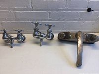 Wide view of three faucets placed on a white surface against a brick wall. Two vintage-style faucets with dual spouts and cross handles on the left, and one modern aged faucet with a single lever handle on the right.