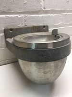Closed view of the metal outdoor wall ashtray showing its oval bowl and mounting plate against a white brick wall.