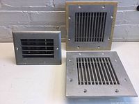 Photo showing all three metal air vents arranged on a surface against a brick wall. Two larger vents have silver finish and one smaller vent is gray.
