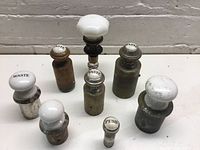 Eight vintage metal and porcelain waste stoppers arranged on white surface against a brick wall background. Various metal finishes with white porcelain tops marked 'WASTE' or 'PUSH'.