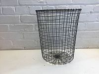Wire waste basket sitting on a flat surface against a white brick wall, showing full side and base view.