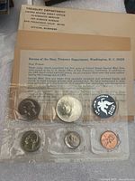 The photo shows the complete set of five proof coins sealed in a transparent plastic sheet along with the original Treasury Department envelope and letter detailing the proof set. The coins visible include the half dollar, quarter, dime, nickel, and penny.