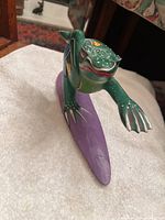 Photo showing front angled view of green wooden frog figurine surfing on purple surfboard with outstretched left arm forward.
