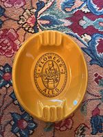 Top-down view of the large oval mustard yellow ceramic ashtray with Flowers Ales logo at center and three cigarette rests visible on one side.
