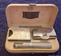 Open ivorene box with GEM logo on lid, showing chrome shaving kit inside: razor handle, razor head with comb, and blade cover.