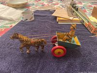 Side view of the entire wind-up cart toy on a blue cloth surface, showing mule, reins, driver, and the cart.