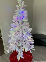 White 6.5 ft artificial Christmas tree with flocked branches, multi-colored pre-lit LED lights turned on, sitting on a red tree skirt.