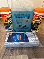 Four cleaning and maintenance products for cars arranged on a wooden surface: one gallon windshield washer fluid in a clear jug, two bottles of Armor All car care products, and a pack of reusable cloths.