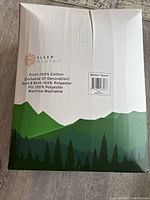 Front side of the box showing Sleep Mantra logo, material composition (front 100% cotton, back and skirt 100% polyester, fill 100% polyester), machine washable, mattress topper label with king size and pure white designation.