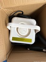 White Grobell steam cleaner in a cardboard box showing top handle and attached power cord with cleaning accessories.