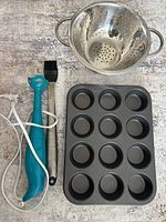 Photo showing all five items: two baking pans (one visible), electric masher, silicone basting brush, and metal strainer.