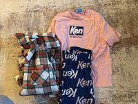 Two men's pajama sets laid out on floor: plaid Christmas-themed pants and peach colored Ken t-shirt with navy blue pants with Ken text