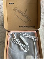 Photo of the lamp and power cords inside the box showing the lamp's LED array, orange trim, and white power cord with switch.