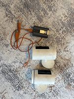 Photo showing two Ring Stick Up Cam Battery security cameras with their mounting bases, two batteries, and one orange USB charging cable on a carpeted floor.