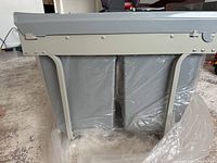 Side view of the metal frame holding two plastic bins wrapped in plastic, showing mounting holes and hinged lid mechanism.