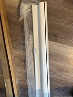 Rolled white fabric window blind with attached pull chain and wrapped hardware, laid on wooden floor.