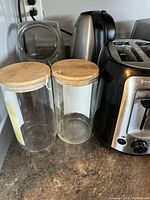 Photo showing two clear glass tea bag holders with wooden lids, positioned next to a toaster and an electric kettle.