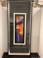 Full view of framed abstract glass artwork showing the vertical rectangular piece with multiple colors in an industrial-style metal frame with visible nick.