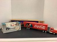 Photo showing both Coca Cola Christmas collector trucks together. The 17 inch semi-truck and trailer is red with Coca Cola branding and Santa imagery, positioned in front of the boxed smaller 1937 Chevrolet truck model in white with Santa.