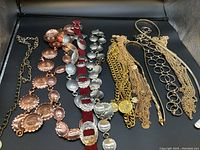 Photo showing various ladies metal belts laid out on black background, including copper conchos, silver oval conchos on red strap, and multiple gold-tone chain link belts with medallions and charms.