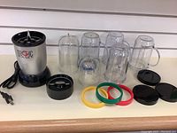 Magic Bullet blender base unit and various jars, colored sealing rings, lids, and blade displayed on a white surface.