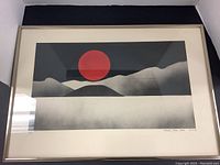 Photo of the framed artwork showing the red sun over mountains in gray scale with the silver frame.