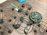 Photo shows six rolling plant stands arranged on floor, five black wire metal designs with wheels, one green decorative floral metal stand with wheels.