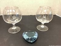 Two clear tulip-shaped brandy glasses and a blue faceted heart-shaped paperweight on a black surface with white background.