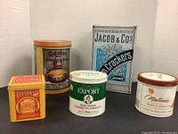 Photo showing five vintage commercial tins including Quaker Corn Meal, Lipton's Tea, Macdonald's Export Cigarette Tobacco, Jacob & Co's Crackers, and Matinée Tabac à Cigarett tins.