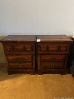 Front view of two matching bedside tables with solid wood construction, decorative carved drawer fronts, and metal handles; visible damage noted on tops and sides