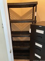 Photo of one black resin shelving unit showing five shelves with grid pattern and vertical supports, positioned next to a black filing cabinet.