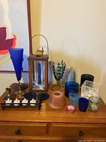 Front view of candles and candlesticks arranged on wooden surface, showing various holders, glass goblet, lantern, blue cone, and candles.