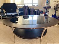 Round coffee table showing top glass inset surrounded by wooden frame with metal legs and wooden bottom shelf in a room.